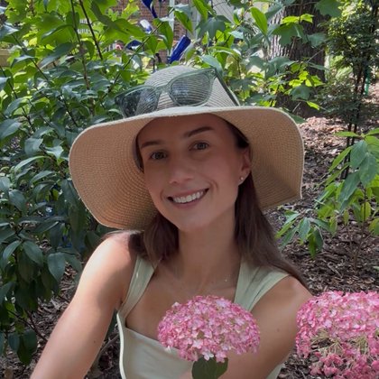 Mia of Mia's Shady Garden, smiling in a wide-brimmed sun hat beside pink hydrangeas